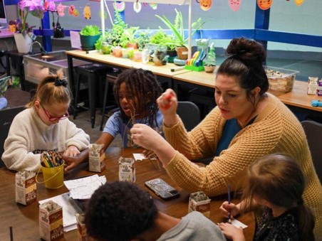 A teacher guides young students in a hands-on activity with cartons and craft supplies in a decorated classroom.