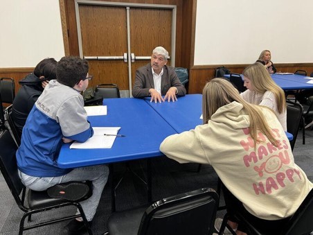 A man speaks with high school students around a blue table as they listen attentively and take notes.