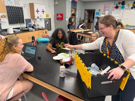 Teacher hands out supplies from a bin to engaged high school students working on a science project.