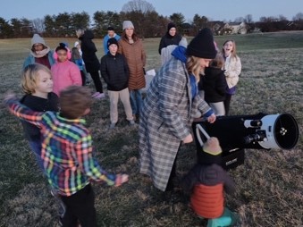At dusk, children and adults in warm clothes gather in a field as a woman helps a child look through a telescope.