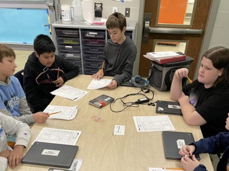 Middle school students sit around a table, working together with pencils and papers while laptops remain closed.