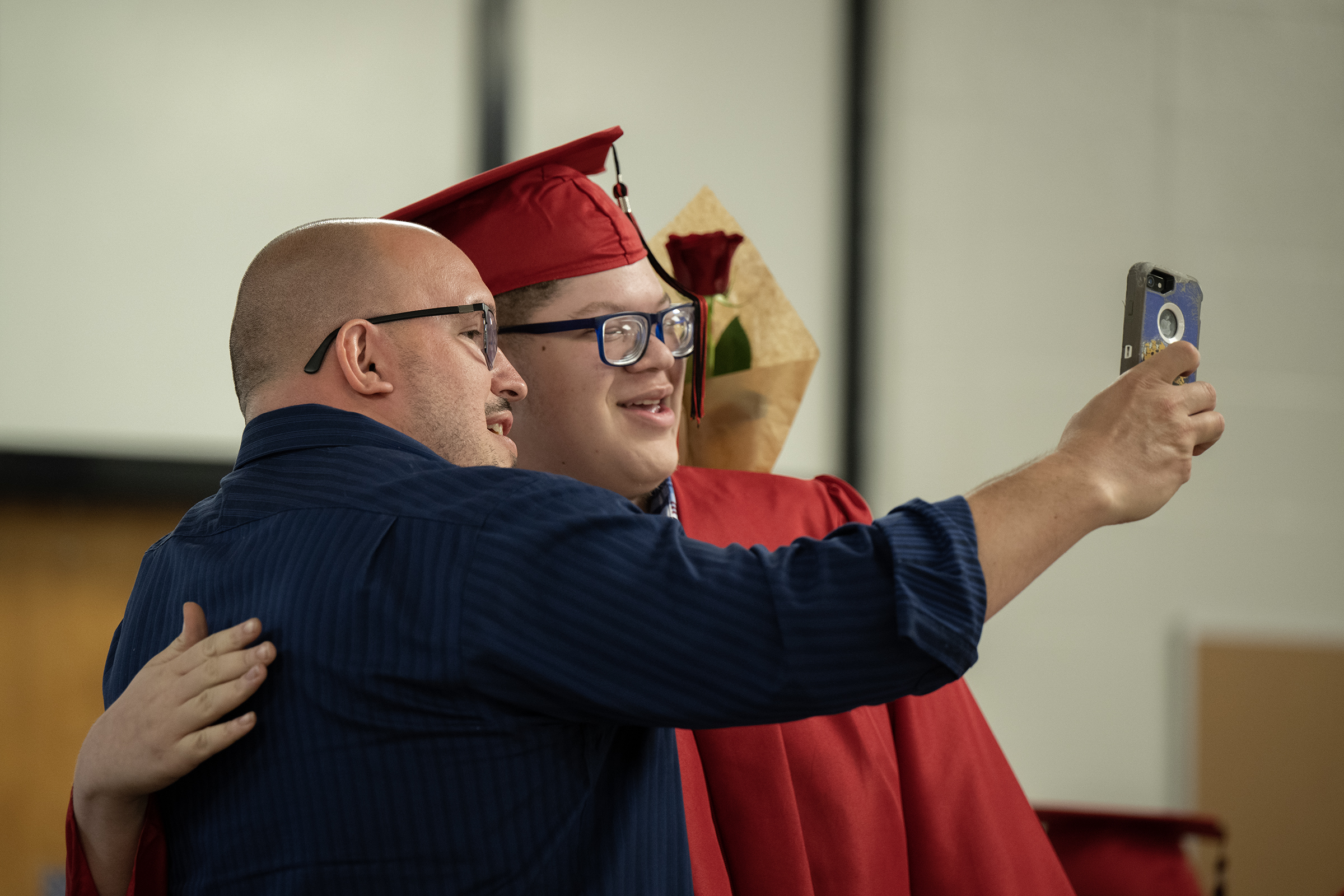 Graduate in red cap and gown taking a selfie with a man during a graduation celebration.